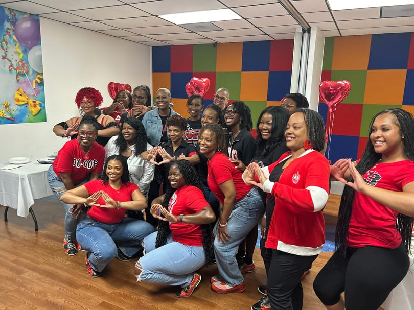 A group of women in red shirts and blue jeans posing with hand gestures and heart balloons.