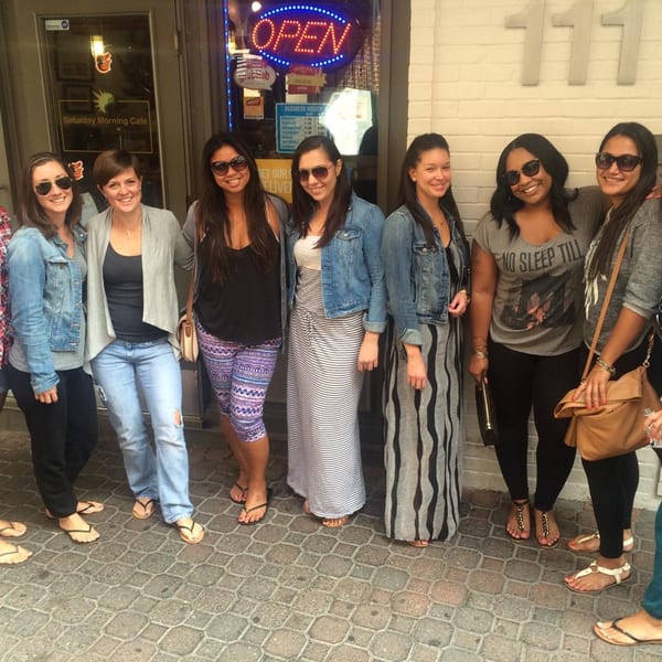 Nine women in casual attire and sunglasses stand smiling outside Saturday Morning Cafe.