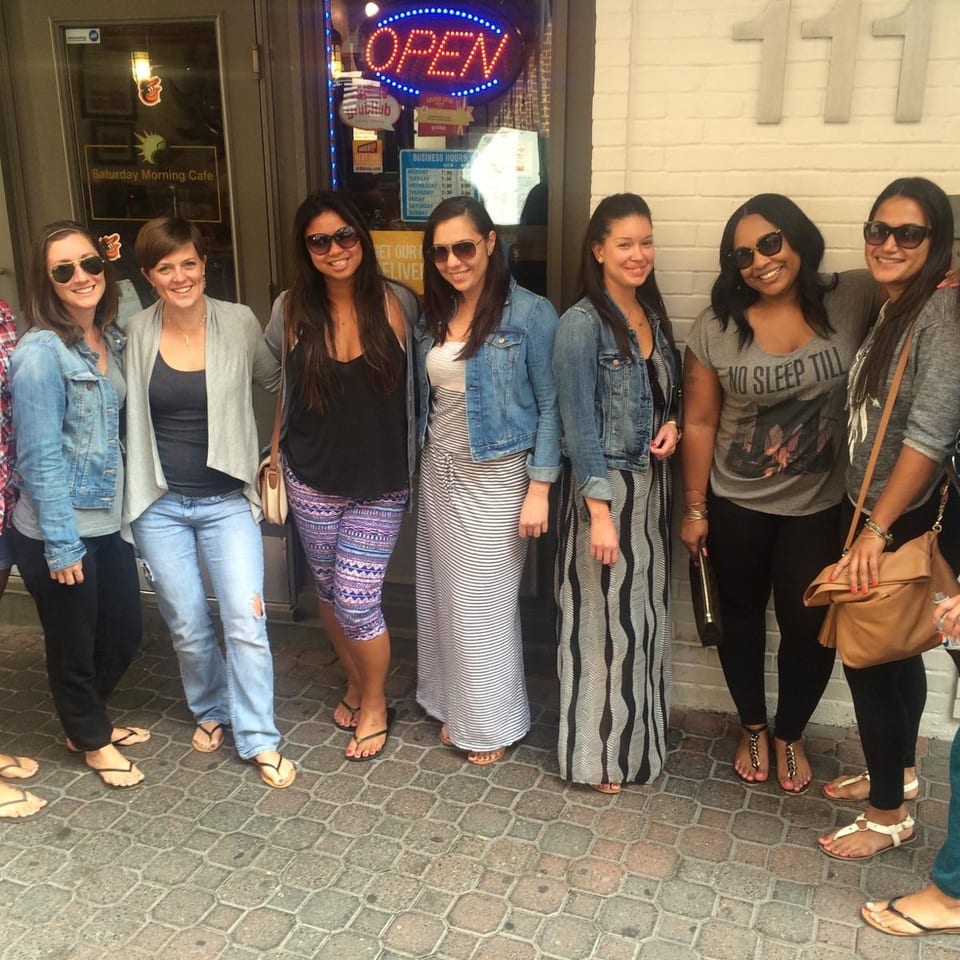 Nine women in casual attire and sunglasses stand smiling outside Saturday Morning Cafe.