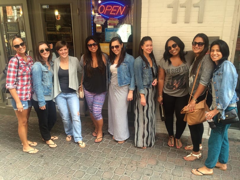 Nine women in casual attire and sunglasses stand smiling outside Saturday Morning Cafe.