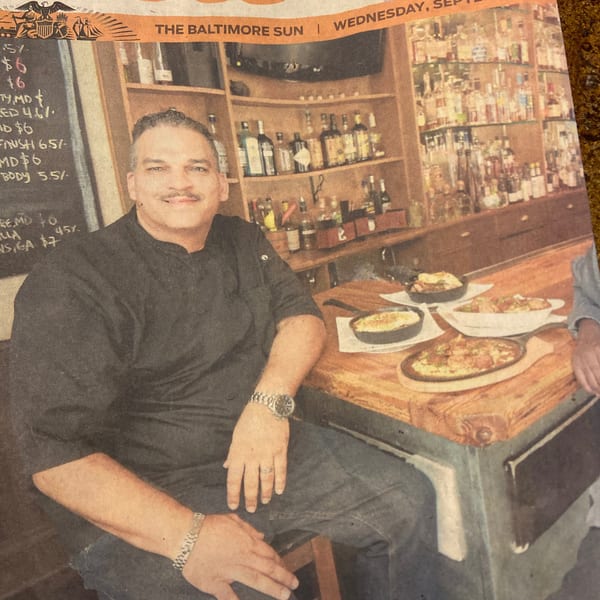 A man in a black chef's coat smiles at a bar with food and drinks.
