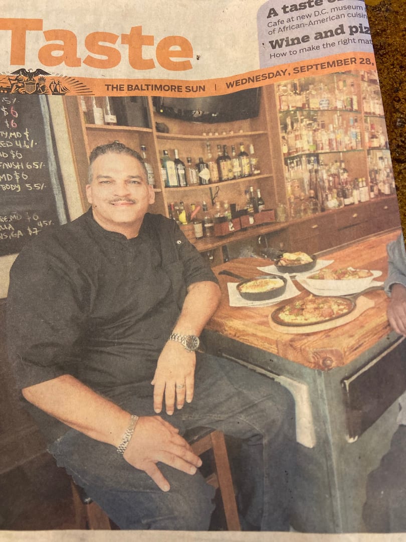 A man in a black chef's coat smiles at a bar with food and drinks.