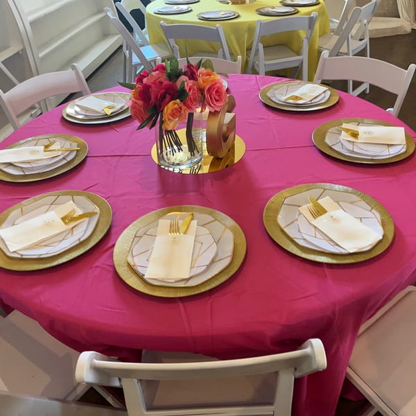 A round table with a hot pink tablecloth, gold chargers, white plates, and a floral centerpiece.