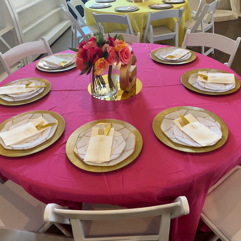 A round table with a hot pink tablecloth, gold chargers, white plates, and a floral centerpiece.