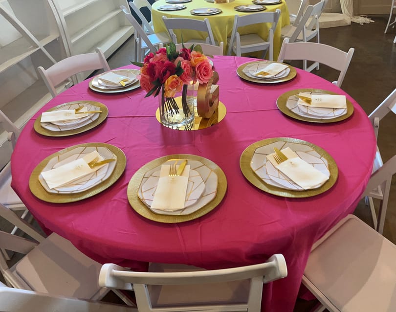 A round table with a hot pink tablecloth, gold chargers, white plates, and a floral centerpiece.