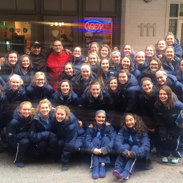 Large group of young women in blue jackets posing with two men in front of a building.