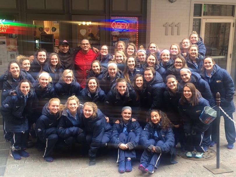 Large group of young women in blue jackets posing with two men in front of a building.