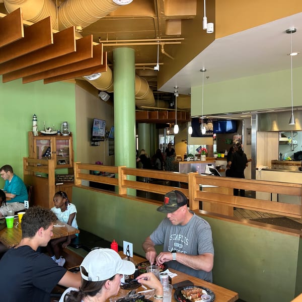 Diners eat at wooden tables in a casual restaurant with light green walls and an open kitchen.