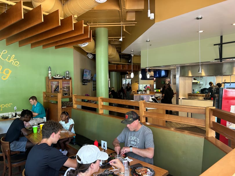 Diners eat at wooden tables in a casual restaurant with light green walls and an open kitchen.