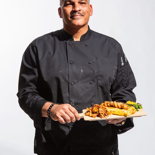 Chef in black uniform holding a wooden board with shrimp, fries, and an omelet.