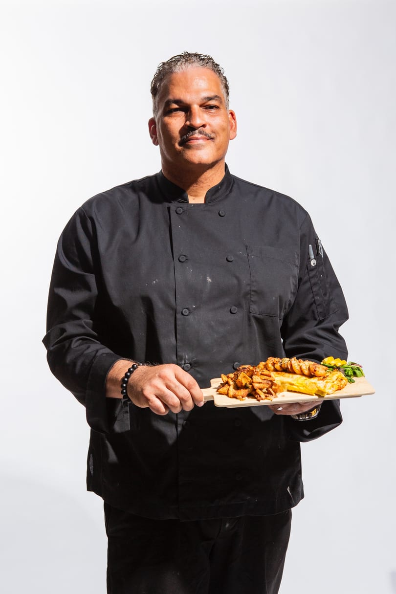 Chef in black uniform holding a wooden board with shrimp, fries, and an omelet.