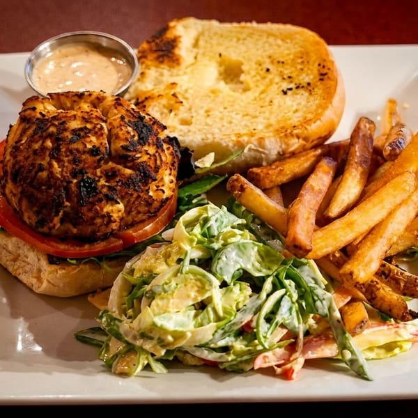 A grilled chicken sandwich with tomato, arugula, fries, coleslaw, and dipping sauce on a white plate.