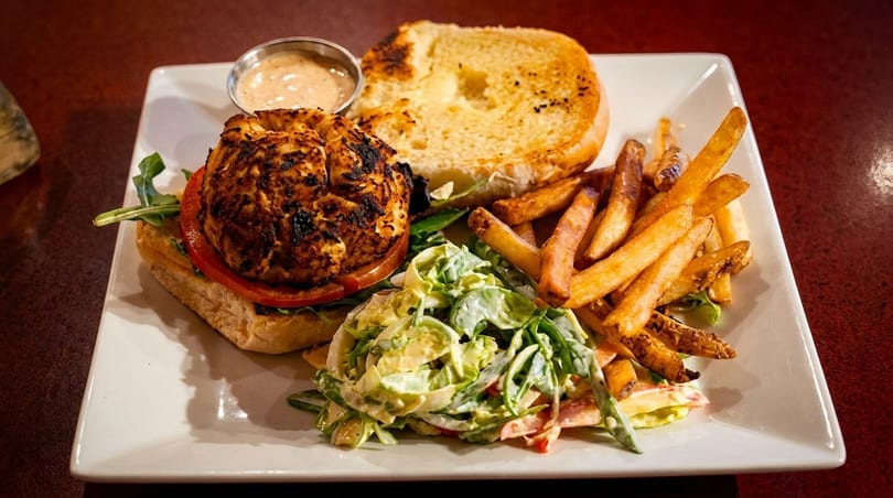 A grilled chicken sandwich with tomato, arugula, fries, coleslaw, and dipping sauce on a white plate.