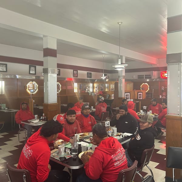 A group of men in red shirts and hoodies are eating at tables in a diner.