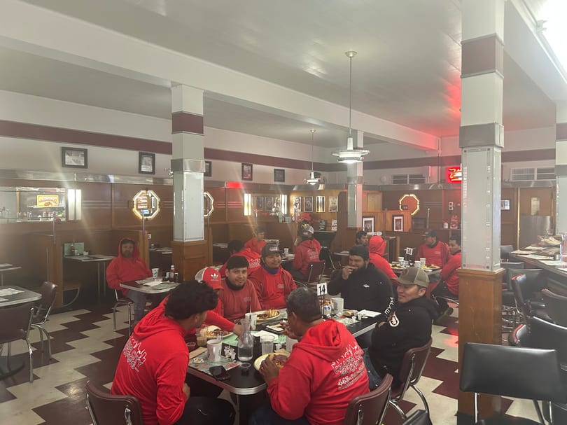 A group of men in red shirts and hoodies are eating at tables in a diner.