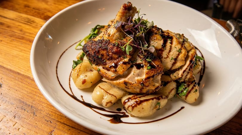Grilled chicken and gnocchi drizzled with balsamic glaze, garnished with microgreens, on a white plate.