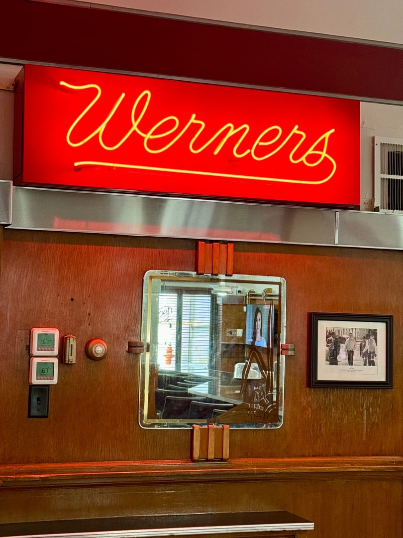 Red neon sign for "Werners" above a wooden wall with a mirror and framed picture.