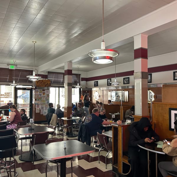 Diners seated at tables and booths in a retro-style restaurant with checkered floors and red accents.