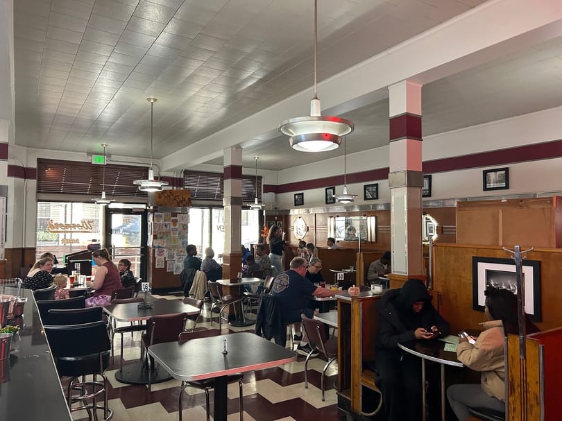 Diners seated at tables and booths in a retro-style restaurant with checkered floors and red accents.