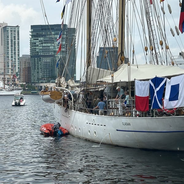 A large white sailboat named "GLADAN" with flags docked in a harbor with city buildings in the background.