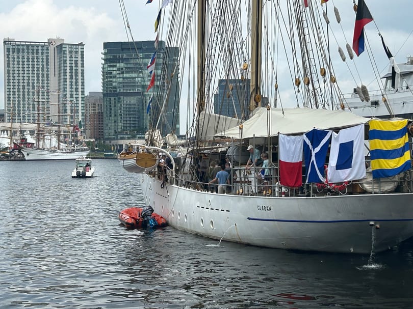 A large white sailboat named "GLADAN" with flags docked in a harbor with city buildings in the background.
