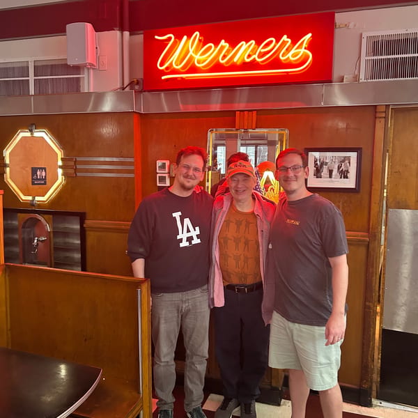 Three men smiling in front of a red neon "Werners" sign inside a diner.