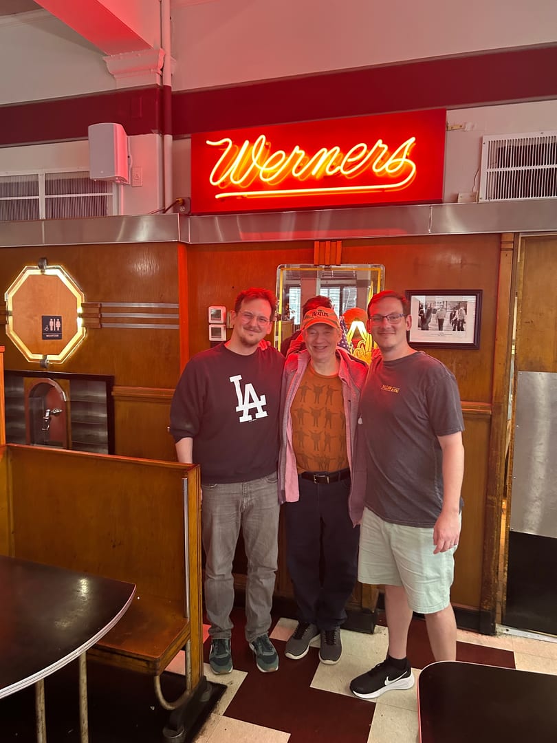 Three men smiling in front of a red neon "Werners" sign inside a diner.