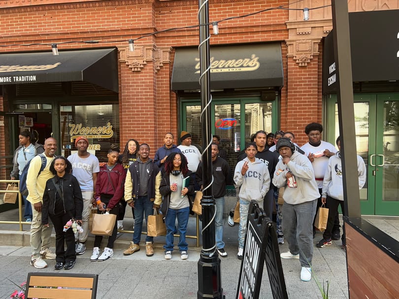 A group of young people and adults stand outside Werner's restaurant, holding brown paper bags.