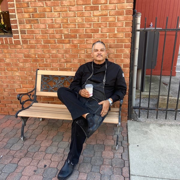 A chef in a black uniform and apron sits on a bench, holding a cup, against a brick wall.