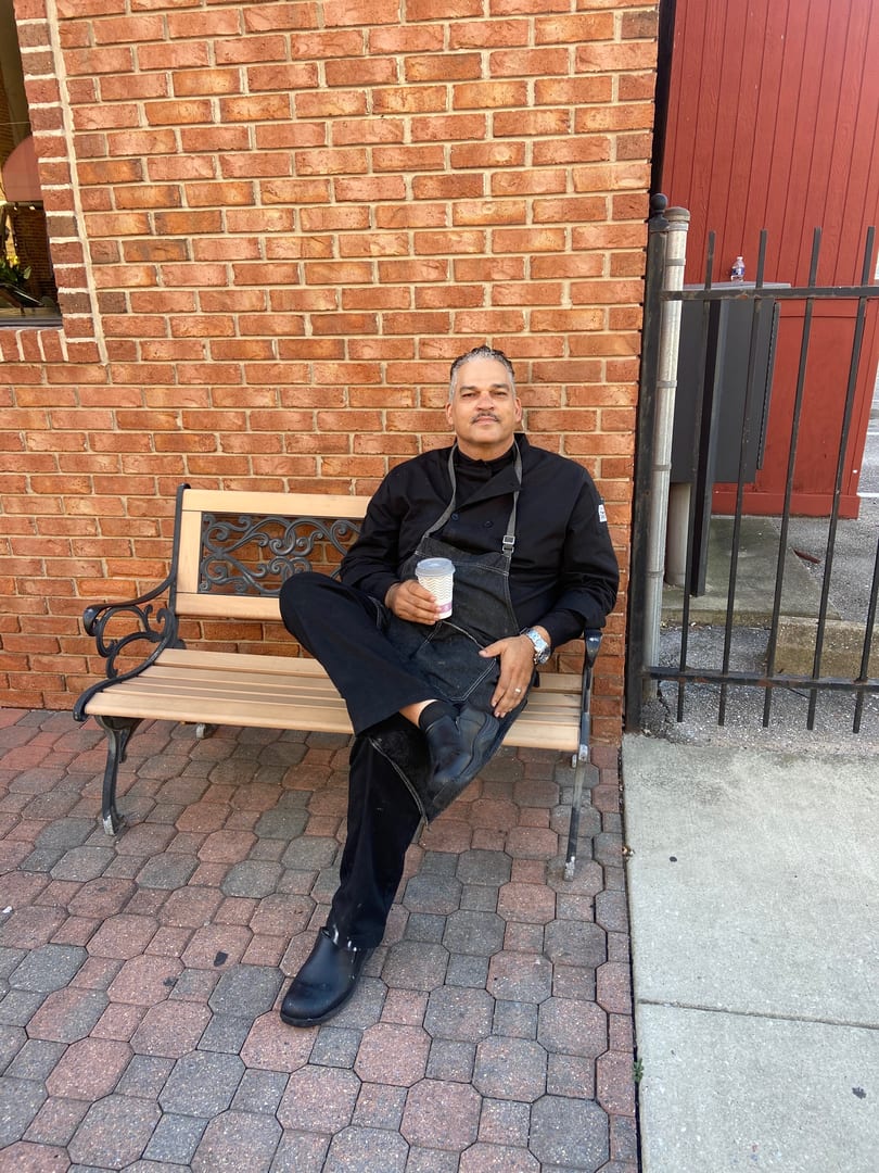 A chef in a black uniform and apron sits on a bench, holding a cup, against a brick wall.