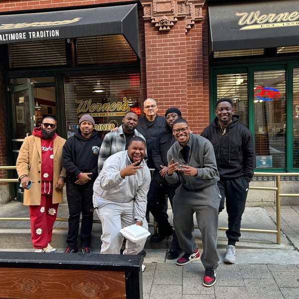 Eight men pose and smile outside Werner's Diner, a brick building with black awnings.