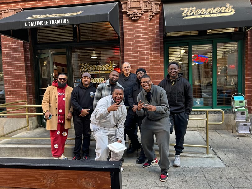 Eight men pose and smile outside Werner's Diner, a brick building with black awnings.