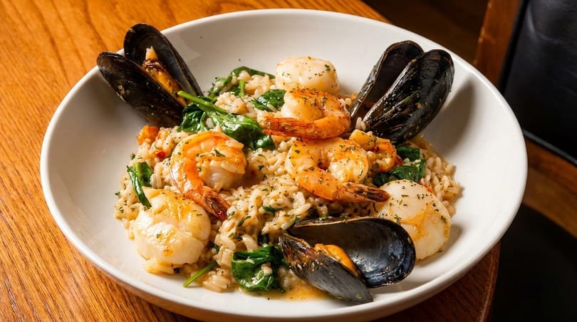Seafood risotto with shrimp, scallops, mussels, and spinach in a white bowl on a wooden table.