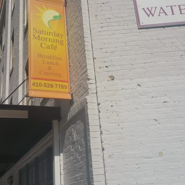 Orange banner for "Saturday Morning Cafe" on a brick building, advertising breakfast, lunch, and catering.