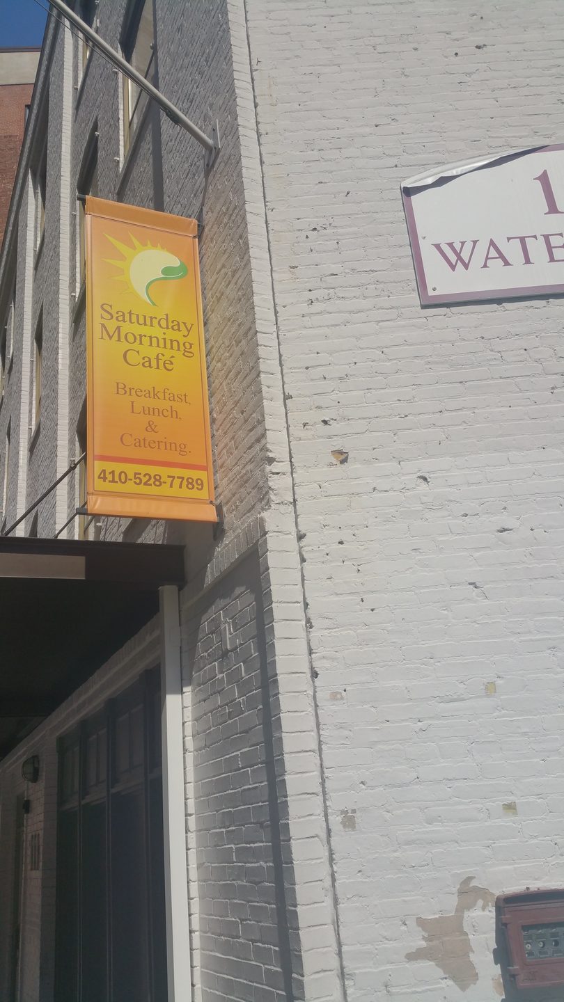 Orange banner for "Saturday Morning Cafe" on a brick building, advertising breakfast, lunch, and catering.