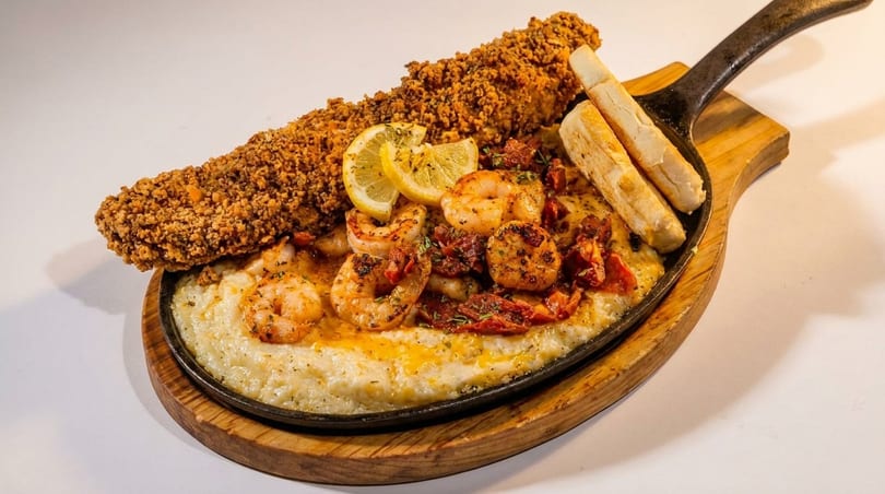 Fried fish and shrimp and grits with lemon slices and breadsticks in a skillet.